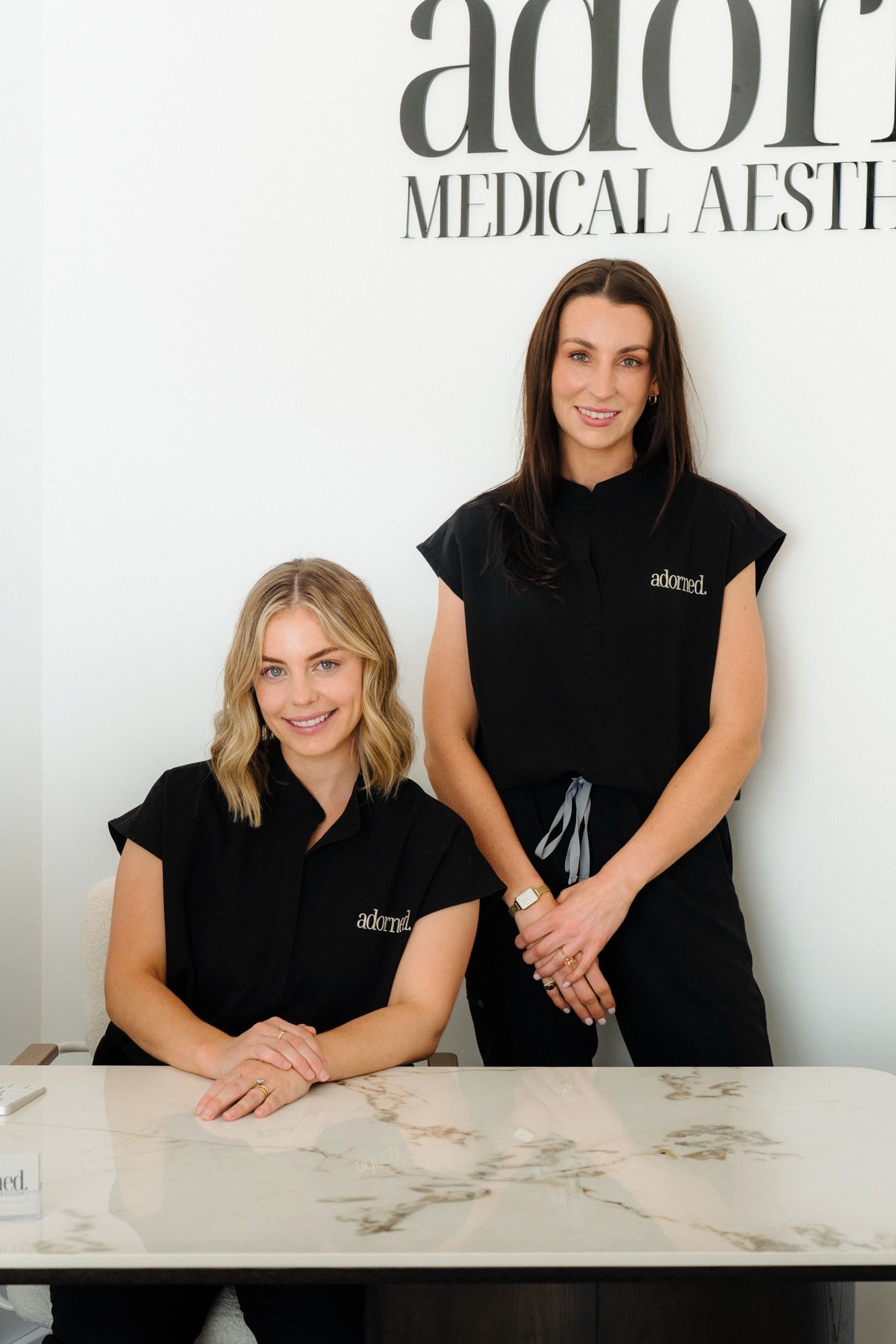 Two women in black 'adormed' shirts standing behind a marble counter.